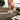 A person in white sneakers, with orange detailing, walking downstairs on a sunny day. The background features green bushes and a clear sky.