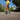 A person runs on a sunny path wearing bright yellow shorts and black sneakers. The scene is surrounded by greenery and houses, conveying an active, vibrant mood.