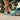 A person wearing bright blue socks stretches their leg beside a pair of gray sneakers on a path. Green trees line the rustic wooden fence.